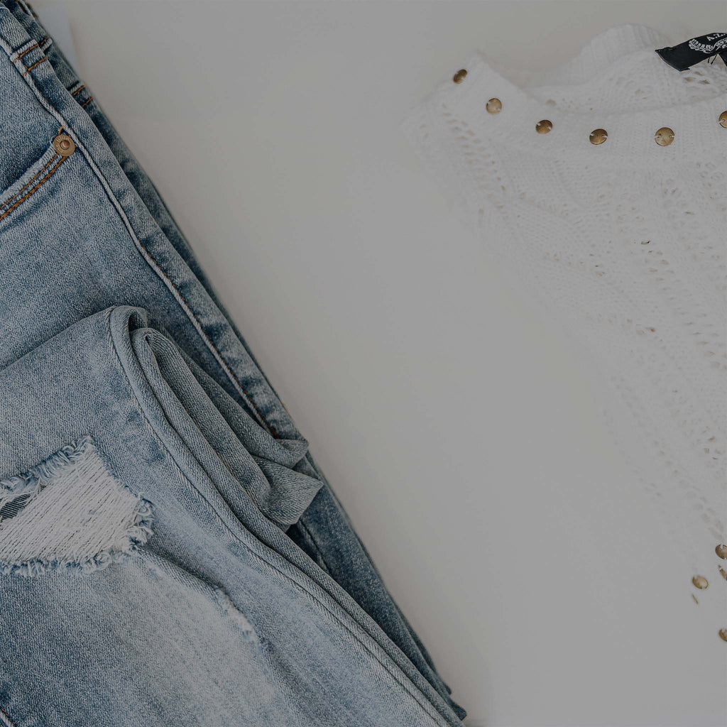 Women's jeans with acid wash and light distressing sitting on a white table top with a white and gold studded sweater